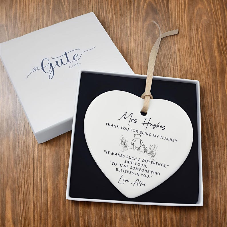 May include: A white heart-shaped ornament with a beige ribbon, presented in a white gift box. The ornament features a Winnie the Pooh illustration and text that reads "Mrs. Hughes, Thank you for being my teacher."