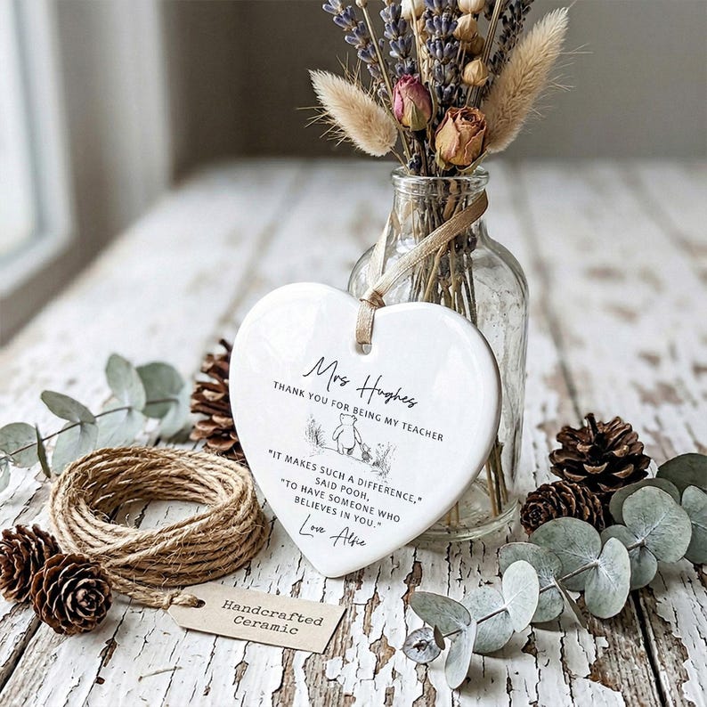 May include: A white heart-shaped ceramic plaque with a ribbon for hanging. The plaque has text that reads "Mrs. Hughes, Thank you for being my teacher." and a Winnie the Pooh illustration. A small tag reads "Handcrafted Ceramic."