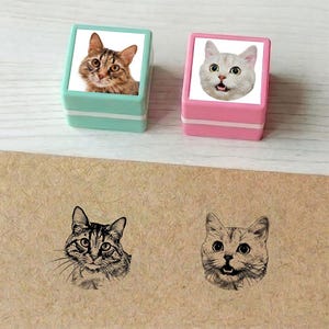 May include: Two cat-themed stampers, one mint green and one pink, each with a cat face. Below, two cat face stamp impressions are visible on a tan surface. The stamps and impressions are in a cat theme.