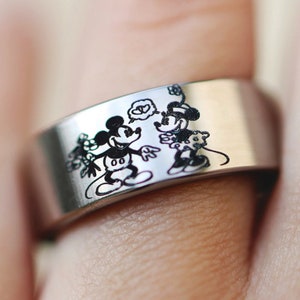 Puede incluir: Un anillo de plata con una imagen grabada en blanco y negro de Mickey Mouse y Minnie Mouse.