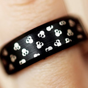 Puede incluir: Un anillo negro con acabado mate, adornado con un patr&oacute;n repetido de peque&ntilde;os dise&ntilde;os blancos estilizados de flores o calaveras. El anillo se muestra en un dedo, destacando los detalles y la textura del anillo.