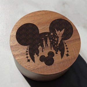 May include: A wooden round box with a laser-engraved design of Mickey Mouse's head with Cinderella's castle and Tinkerbell flying above the castle. The design is in black and the box is a light brown color.