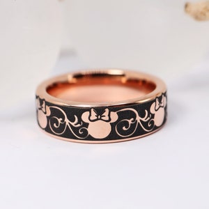 Op de afbeelding: Een zwarte ring met een ros&eacute;gouden band met een herhalend patroon van Minnie Mouse-hoofden met een strik.