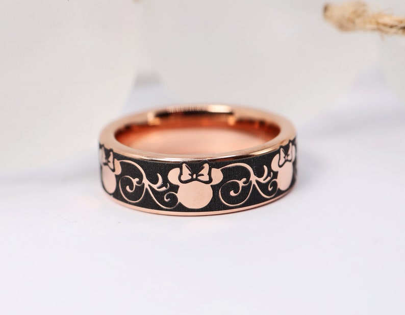 Op de afbeelding: Een zwarte ring met een ros&eacute;gouden band met een herhalend patroon van Minnie Mouse-hoofden met een strik.