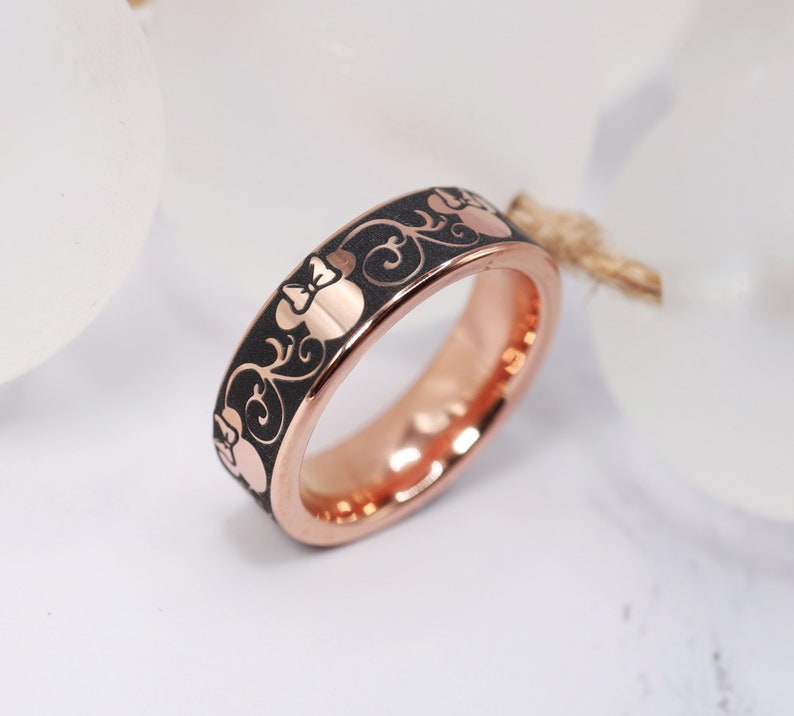 Op de afbeelding: Een ros&eacute;gouden ring met een zwarte emaille afwerking en een herhalend patroon van een cartoon muizenkop met een strik.