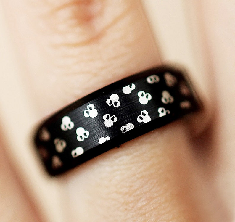 Puede incluir: Un anillo negro con acabado mate, adornado con un patr&oacute;n repetido de peque&ntilde;os dise&ntilde;os blancos estilizados de flores o calaveras. El anillo se muestra en un dedo, destacando los detalles y la textura del anillo.