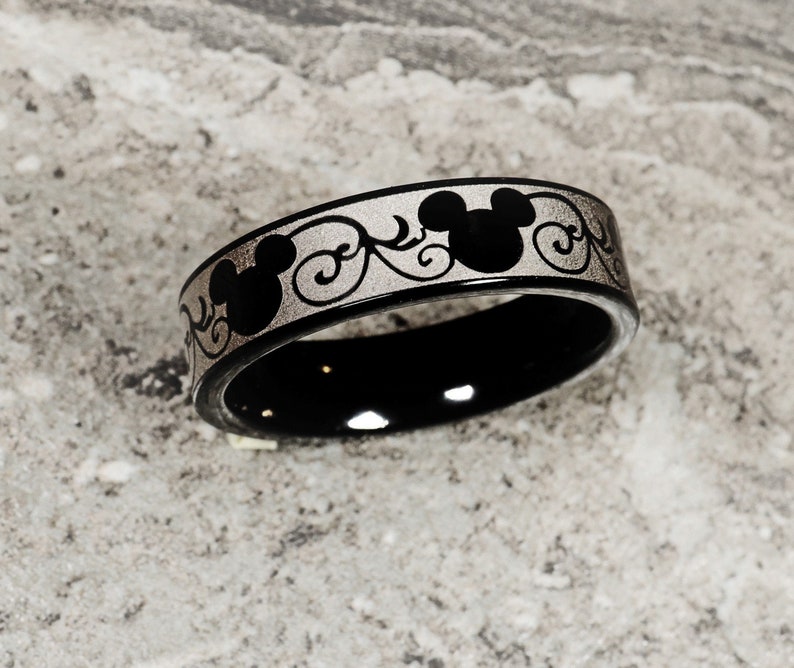 May include: Black ceramic ring with a silver band featuring a repeating pattern of Mickey Mouse heads and swirls.