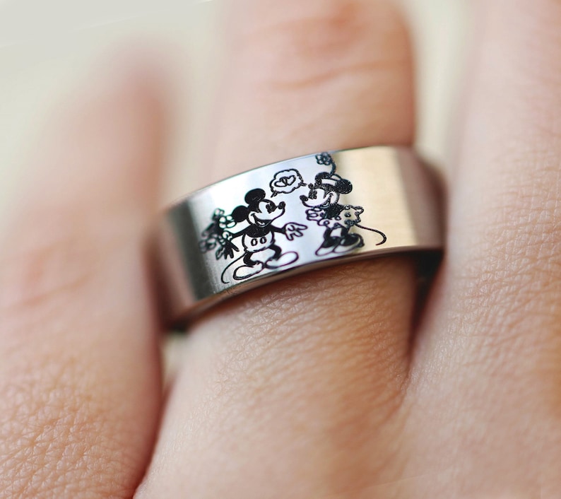 Puede incluir: Un anillo de plata con una imagen grabada en blanco y negro de Mickey Mouse y Minnie Mouse.