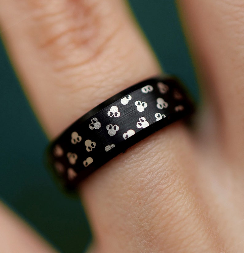 Puede incluir: Un anillo negro con acabado mate, adornado con un patr&oacute;n de peque&ntilde;os dise&ntilde;os de calaveras plateadas. El anillo se muestra en un dedo, con una vista de cerca que resalta los detalles de la joya.