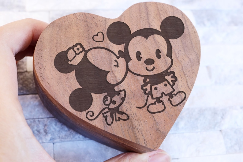 May include: A wooden heart-shaped box with a laser-engraved design of Mickey Mouse and Minnie Mouse kissing. The box is made of dark wood and has a smooth finish.