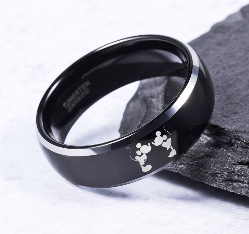 May include: Black tungsten carbide ring with a silver beveled edge and a laser engraved image of Mickey Mouse and Minnie Mouse holding hands. The ring is engraved with the words "Tungsten Carbide".