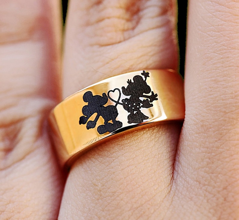 May include: A gold ring with a black silhouette of Mickey Mouse and Minnie Mouse holding hands with a heart between them.