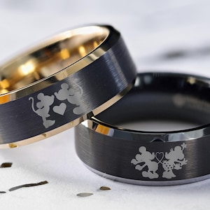 May include: Two black tungsten carbide wedding bands with a gold inlay. The bands feature an engraved design of Mickey Mouse and Minnie Mouse holding hands with a heart.