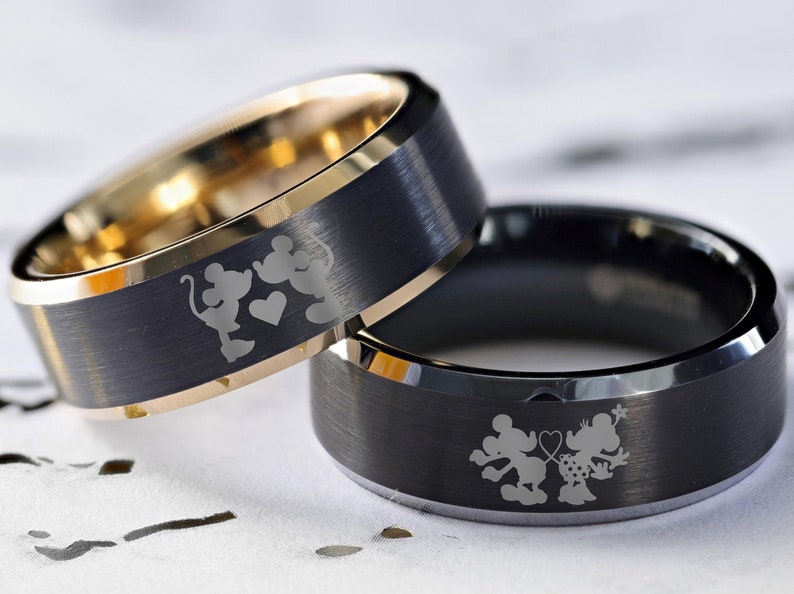 May include: Two black tungsten carbide wedding bands with a gold inlay. The bands feature an engraved design of Mickey Mouse and Minnie Mouse holding hands with a heart.