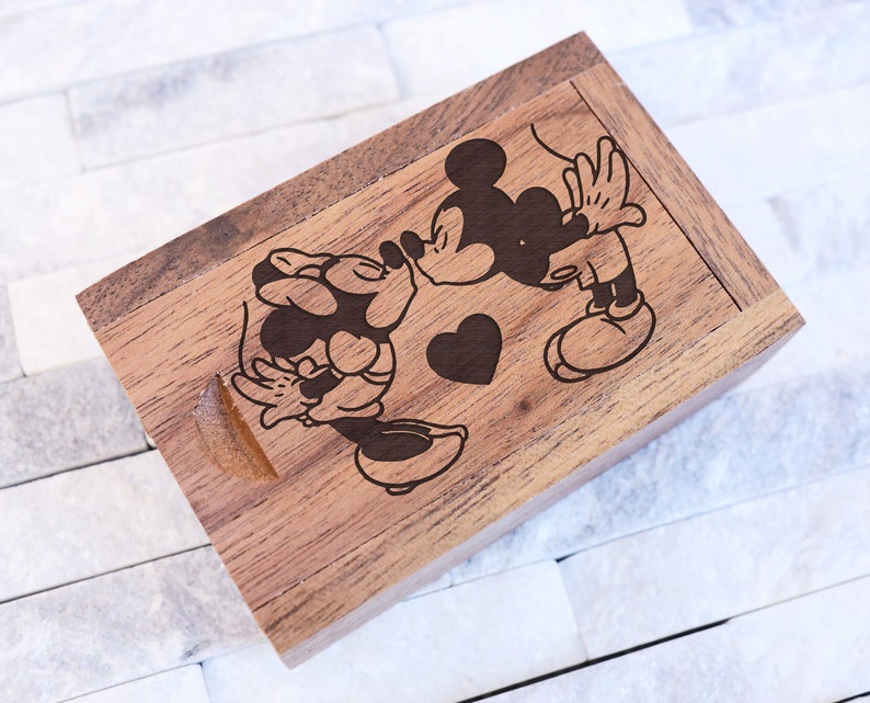 May include: A wooden box with a laser engraved design of Mickey Mouse and Minnie Mouse kissing. The box is brown and has a heart shape in the center.