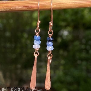 May include: Handmade earrings with copper wire, teardrop pendants, and blue gemstone beads. The earrings have copper ear wires and feature a series of blue and light blue beads. The website address bluemoonglade.com is visible at the bottom of the image.