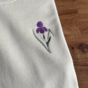 May include: A light beige sweatshirt with a purple and green embroidered iris flower on the front.