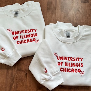 May include: Two white sweatshirts with red embroidered hearts and the text "University of Illinois Chicago" on the front. The sweatshirt on the left has a "C" embroidered on the left sleeve and the sweatshirt on the right has a "J" embroidered on the left sleeve.