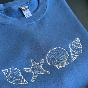 May include: A blue sweatshirt with white embroidered seashells and a starfish. The sweatshirt is folded in half.