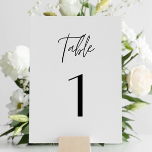 May include: White card with black text that reads "Table 1" on a wooden stand. The card is in front of a bouquet of white flowers.
