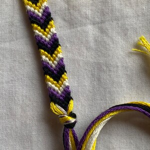 May include: A friendship bracelet with a chevron pattern in yellow, white, purple, and black colors.