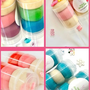 May include: Four clear plastic tubes filled with colorful play dough. Each tube has a different color combination and is topped with various decorative elements, such as a butterfly, a snowman, and a snowflake. The tubes are labeled "littlest dough shop".