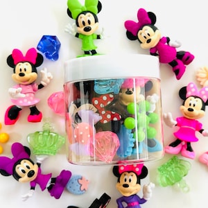 May include: A clear plastic jar filled with colorful plastic charms and figurines, including Mickey Mouse characters. The jar is surrounded by a variety of colorful plastic charms and figurines, including Mickey Mouse characters, crowns, and diamonds.