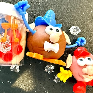 May include: A playful set of Mr. Potato Head-style clay figures with various colorful accessories. The set includes a clear container filled with additional parts, and several clear, faceted plastic gems. The figures are brown and pink with blue, yellow, and red accents.