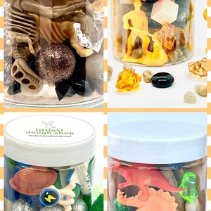May include: Four clear plastic jars with white lids filled with various small toys and objects. The jars are labeled "littlest dough shop" and "littlestdoughshop.com". The jars contain a dinosaur, a construction worker, a skeleton, and an astronaut.