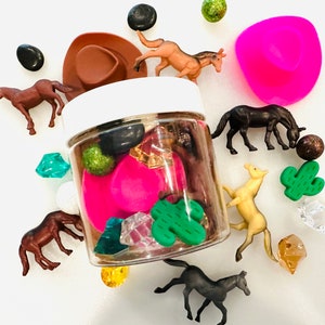May include: A clear glass jar filled with various small objects, including toy horses, a pink cowboy hat, green cactus, and colorful gems. The jar is sitting on a white surface.