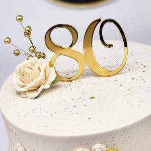 May include: A white frosted cake decorated with gold accents, including a '80' cake topper and floral embellishments. The cake is adorned with a gold number 80, a cream-colored rose, and gold-colored decorative sprigs.