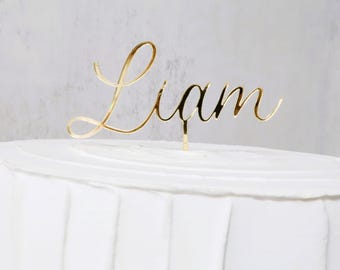 Può includere: Un cake topper dorato con il nome "Liam" in corsivo. Il topper è inserito in una torta glassata bianca. Lo sfondo è di colore grigio chiaro.