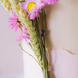 May include: A bouquet of dried flowers with pink and white blossoms and wheat stalks. The flowers are tied together with a clear elastic band.