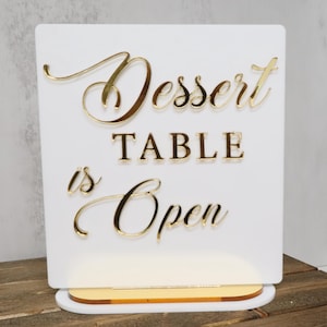 Op de afbeelding: Een wit bord met gouden letters met de tekst "Dessert TABLE is Open". Het bord heeft een gouden voet en is ontworpen om rechtop te staan. Het bord is waarschijnlijk voor een bruiloft of feest.