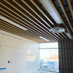 May include: A modern office space with a wood slat ceiling and a large window. The ceiling has a circular air vent and a white wall with a light switch.