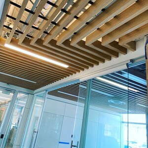 May include: A modern office space with a wood slat ceiling and glass walls. The ceiling is made of light brown wood slats arranged in a grid pattern. The walls are made of clear glass panels.