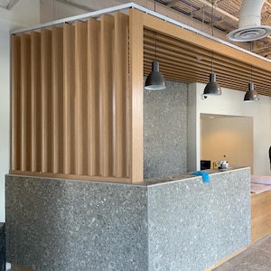 May include: A modern office reception desk with a gray speckled countertop and a wood slat ceiling. The desk is situated in front of a wall with a gray speckled finish. Three black pendant lights hang from the ceiling.