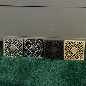 Könnte beinhalten: Vier quadratische Abflussabdeckungen in verschiedenen Ausführungen: Silber, Schwarz und Gold. Jede Abdeckung hat ein geometrisches Design.
