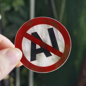 Op de afbeelding: Een ronde sticker met een witte achtergrond en de zwarte tekst AI in het midden. Een dikke rode cirkelvormige rand omringt de tekst en een diagonale rode lijn loopt door het midden, wat een verbodsteken tegen kunstmatige intelligentie aangeeft.