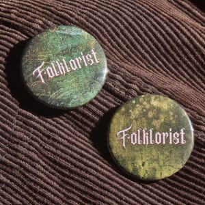 May include: Two round green and gold buttons with the word "Folkloreist" in a decorative font. The buttons are on a brown corduroy fabric.