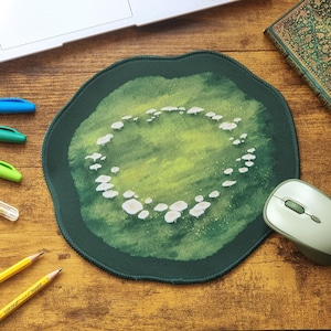 Può includere: Un tappetino per mouse rotondo con bordo verde scuro e un'illustrazione centrale di piccoli funghi bianchi disposti in un motivo circolare su uno sfondo strutturato con sfumature di verde chiaro e scuro, creando un effetto pittorico.