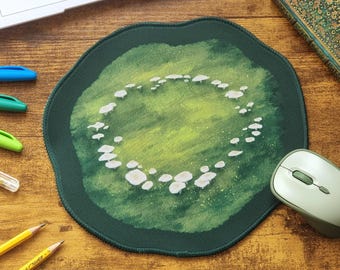 Op de afbeelding: Een ronde muismat met een donkergroene rand en een centrale illustratie van kleine witte paddenstoelen in een cirkelvormig patroon tegen een textuurrijke achtergrond van lichte en donkere groentinten voor een zacht effect.