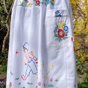 May include: White apron with embroidered floral and pastoral scenes. Features a pocket with floral embroidery and a design of a child running with a goose. The apron has a pink ribbon tie at the waist.