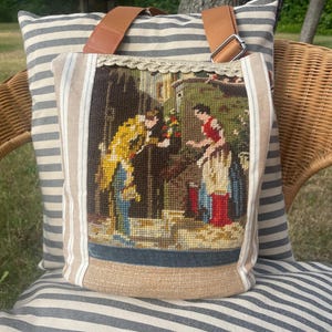 Può includere: Una borsa tote con un pannello a mezzo punto che raffigura due figure in un giardino. La borsa presenta lati in tessuto color cuoio con strisce bianche, cinghie marroni regolabili e un bordo intrecciato decorativo sulla parte superiore.