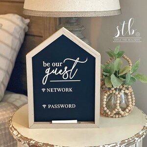 May include: A white wooden sign shaped like a house with a black chalkboard insert. The sign says "be our guest" in white cursive lettering. Below the text are two icons for wifi network and password. The sign is sitting on a white side table with a small plant in a glass vase.