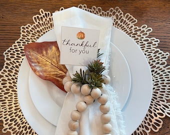 Puede incluir: Una mesa con temática otoñal con un plato blanco, una servilleta y un servilletero de cuentas de madera. Una tarjeta con una ilustración de calabaza y el texto "thankful for you" se coloca en la servilleta. Una hoja decorativa y un mantel individual dorado completan el arreglo.