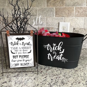 May include: Black metal bucket with a white "trick or treat" sign and a spider web design. A sign with the text "trick or treat take a sweet two or three if you dare but please leave some to spare! Happy Halloween!" is in front of the bucket.