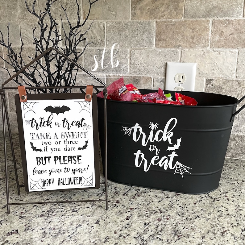 May include: Black metal bucket with a white "trick or treat" sign and a spider web design. A sign with the text "trick or treat take a sweet two or three if you dare but please leave some to spare! Happy Halloween!" is in front of the bucket.
