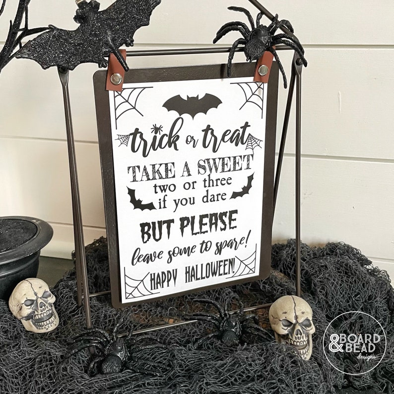 May include: Black and white printable sign with the text "trick or treat take a sweet two or three if you dare but please leave some to spare! happy halloween!"  The sign is on a black metal easel with a spider and bat decoration.