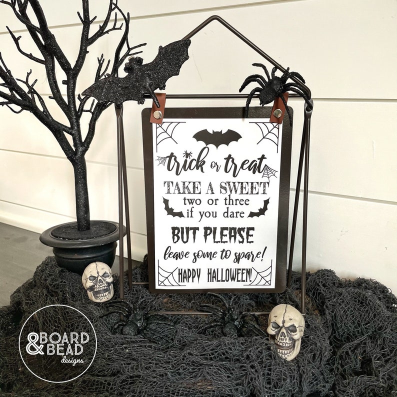 May include: Black and white printable sign with the text "trick or treat take a sweet two or three if you dare but please leave some to spare! happy halloween!" on a black background with a spider web design. The sign is displayed on a black metal easel with a black spider and bat decorations.